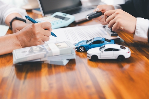 Close-up of two people reviewing auto loan paperwork at a table, with a calculator, a stack of cash, and two small toy cars—one blue and one white—representing vehicle financing