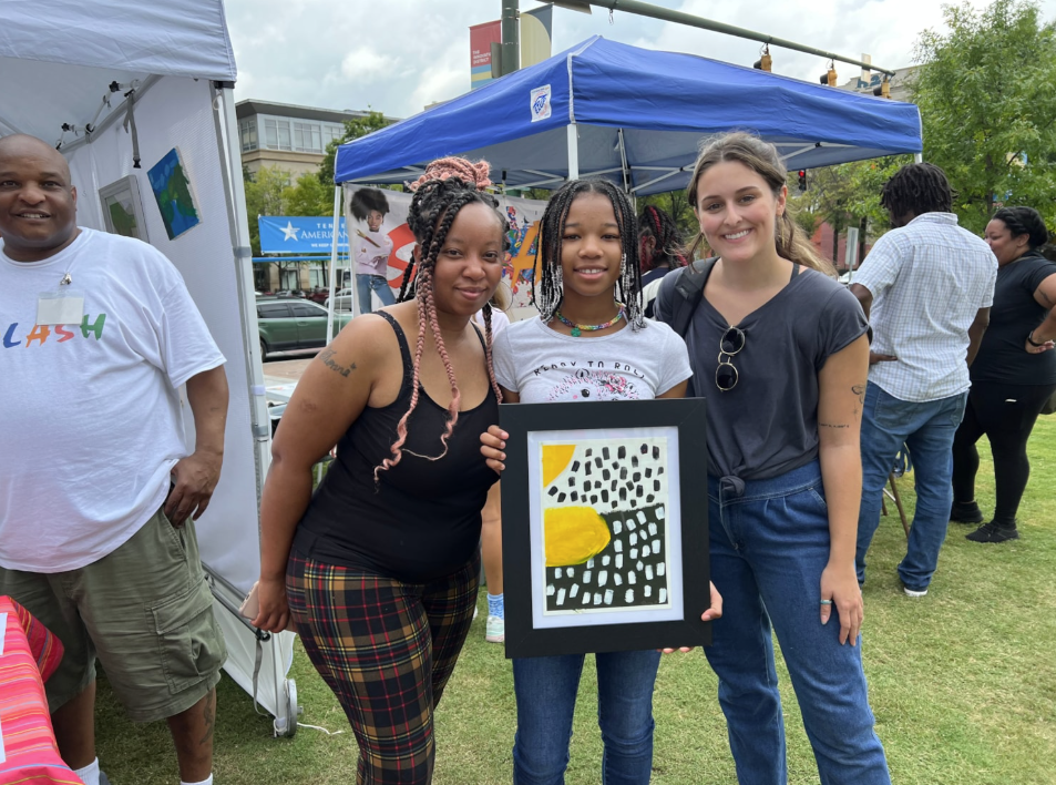 A girl and two young women standing in a park holding a piece of artwork.